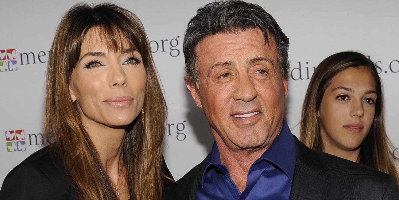 Vi ser Jennifer Flavin till vänster, hon bär en svart kavaj. Till höger ser vi Sylvester Stallone i en blå skjorta och svart kostym, de bägge ler.