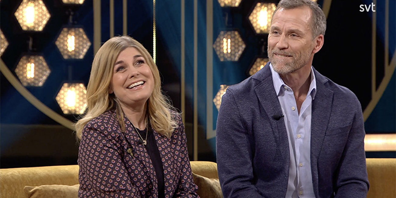 Till vänster ser vi en glad Pernilla Wahlgren och till höger ser vi en glad Christian Bauer, de sitter i en tv-soffa.